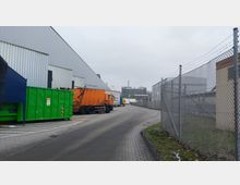 Industriegelände mit gepflastertem Weg, ergänzt durch große, metallverkleidete Gebäudewände auf der linken Seite. Im Vordergrund befinden sich ein grüner Container und ein orangefarbener Müllwagen, rechts ein Maschendrahtzaun, im Hintergrund sind weitere Gebäude und Schornsteine sichtbar.