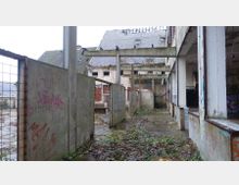 Verlassenes Industriegelände mit beschädigten Gebäuden, überwuchertem Boden und Graffiti an den Wänden. Teile der Konstruktion, wie Stahlträger und offene Bereiche, sowie ein eingestürztes Dach sind sichtbar.