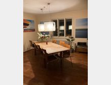 Esszimmer mit einem langen Holztisch und acht passenden Holzstühlen. Eine große, rechteckige Hängelampe mit segmentierter Struktur hängt über dem Tisch, umgeben von dekorativen Bildern an den Wänden, einem Fenster und Pflanzen.