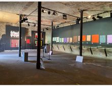 Ein industriell anmutender Ausstellungsraum mit unverputzten Ziegeldecken und Wänden, in dem moderne Kunstwerke an schwarzen Wandflächen hängen. Beleuchtungsscheinwerfer sind an Deckenstangen befestigt, und einzelne Kleidungsstücke sind an einem Kleiderständer ausgestellt.