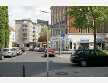 Straßenecke mit Wegweisern für „Bayerischer Platz“ und „Innsbrucker Straße“, umgeben von mehrstöckigen Wohn- und Geschäftsgebäuden, darunter ein rotes Backsteinhaus mit Geschäften im Erdgeschoss. Geparkte und fahrende Autos sowie einige Passanten sind sichtbar, daneben stehen Bäume am Straßenrand.