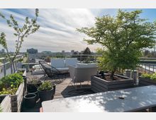 Eine moderne Dachterrasse mit Holzboden, weißen Sitzmöbeln, Blumentöpfen und Hochbeeten mit Pflanzen. Im Hintergrund sind eine Baumreihe und eine städtische Skyline unter einem leicht bewölkten Himmel zu sehen.