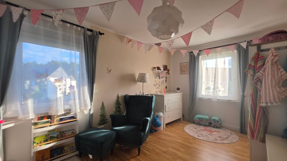 Helles Kinderzimmer mit zwei großen Fenstern, grauen Vorhängen und einer Wimpelgirlande an der Decke. Im Raum stehen ein Lesesessel mit Hocker, ein niedriges Bücherregal, eine weiße Kommode sowie Spielzeug und ein runder Teppich auf hellem Holzboden.