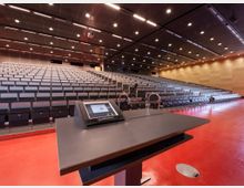 Moderner Hörsaal mit gestuft angeordneten Sitzreihen und einer Rednertribüne im Vordergrund, ausgestattet mit einem Bedientablet und Mikrofonen. Der Raum hat eine rote Bodenfläche, eine Holzverkleidung an den Wänden und eine mit Einbauleuchten versehene Decke.
