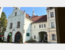 Historisches Gebäude mit weißer Fassade, roten Ziegeldächern und Rundbogenfenstern, in dessen Innenhof ein überdachter Durchgang führt; im Hintergrund ist der Berliner Fernsehturm sichtbar. Auf der linken Seite erkennt man Pflanzen und ein Schild eines Restaurants.