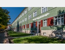 Wohngebäude mit grün verputzter Fassade und weißen Fenstern, akzentuiert durch rot gestrichene Fensterläden; davor eine gepflegte Rasenfläche und ein gepflasterter Gehweg entlang der Straße mit Bäumen.