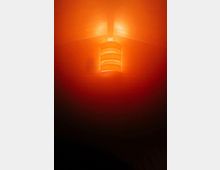 Innenansicht einer hölzernen Decke mit zentralem Deckenbalken, an dem eine zylindrische Deckenleuchte mit mehreren Lamellen hängt. Die Leuchte strahlt ein warmes orangefarbenes Licht aus und beleuchtet den Deckenbereich.