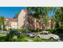 Straßenansicht in einem Wohngebiet mit mehrstöckigen, hellbraunen Wohnhäusern und roten Ziegeldächern, umgeben von Bäumen und Grünflächen. Im Vordergrund parken mehrere Autos, darunter ein beigefarbener VW Käfer, entlang einer ruhigen Straße.