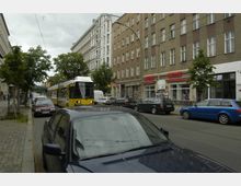 Straßenszene in einer Stadt mit mehrstöckigen Wohn- und Geschäftsgebäuden, einem gelb-weißen Straßenbahnwagen auf den Gleisen, parkenden Autos am Straßenrand und kleinen Bäumen entlang des Gehwegs.