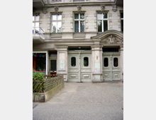 Fassade eines historischen Mehrfamilienhauses mit verzierten Elementen über den Fenstern und einer doppelten Eingangstür aus Holz. Links befindet sich ein kleines Geschäft mit Schaufenster, und vor dem Gebäude führt ein gepflasterter Gehweg entlang.