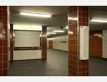 U-Bahn-Station mit rot-weißen gefliesten Wänden und Säulen. Im Vordergrund ein geschlossenes Bistro mit einem Rollladen und ein Fahrplan der Linie U6, im Hintergrund die Beschilderung „Alt-Mariendorf“.