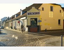 Straßenszene mit Kopfsteinpflaster und einer gelben Gebäudefassade, die eine Bäckerei mit großem Schild zeigt. Ein Radfahrer fährt auf der Straße, die von weiteren niedrigen Gebäuden mit roten und grauen Dächern gesäumt ist.