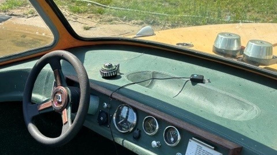 Blick aus dem Innenraum eines Oldtimers auf Armaturenbrett und Lenkrad, mit analogen Rundinstrumenten und einem kleinen Handschuhfach. Durch die gewölbte Frontscheibe sind die Motorhaube sowie ein grüner Garten im Hintergrund zu sehen.