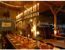 Eine rustikale Scheune mit Holzbalken und Ziegeldach, eingerichtet für ein Abendessen mit einer langen, gedeckten Holztafel. Dekorative Elemente wie hängende Kerzen, Blumenarrangements und Lichterketten schaffen eine stimmungsvolle Atmosphäre.