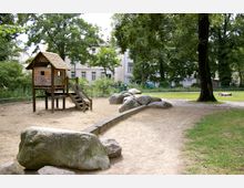 Ein Spielplatz mit einem Holzspielhaus auf Stelzen und einer kleinen Rutsche, umgeben von Sandfläche, Steinen und Rasen. Im Hintergrund sind Bäume, ein Zaun und mehrstöckige Wohnhäuser sichtbar.