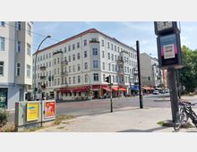 Straßenecke mit einem vierstöckigen Wohngebäude mit heller Fassade und kleinen Balkonen, im Erdgeschoss befinden sich Geschäfte und Cafés mit überdachten Sitzbereichen. Im Vordergrund sind Gehwege, ein Verkehrssignal und eine Litfaßsäule, im Hintergrund Bäume und weitere Gebäude.