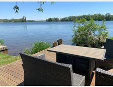 Terrasse mit Holzboden und Rattanmöbeln, bestehend aus einem Tisch und mehreren Stühlen, direkt am Ufer eines Sees. Im Hintergrund sind grasbewachsene Uferbereiche und Bäume unter klarem, blauem Himmel zu sehen.