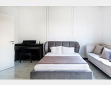 Helles Schlafzimmer mit einem grauen Polsterbett, einem weißen Bettlaken und Kissen. Im Hintergrund steht ein schwarzes Klavier mit einem Hocker, rechts daneben ein helles Sofa mit Kissen.