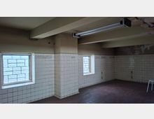 Ein leerer Raum mit weißen, abgenutzten Fliesenwänden bis zur halben Raumhöhe, zwei kleinen Fenstern mit Glasbausteinen und einer Decke mit sichtbaren Trägern und einer Leuchtstofflampe. Der Boden besteht aus dunklen, rötlichen Fliesen.