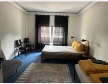 Ein einfach eingerichtetes Schlafzimmer mit blauem Teppichboden, einem Doppelbett mit gelben Kissen und Bettdecke, einem zusätzlichen Bett, einem kleinen runden Tisch mit zwei Stühlen und einem grauen Sofa. Das Zimmer ist durch ein Fenster mit schwarzen Vorhängen beleuchtet, und ein gemusterter Teppich liegt auf dem Boden.