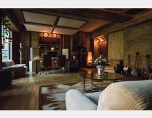 Ein Musikstudio mit Holzwänden und einer warmen Beleuchtung, ausgestattet mit Schreibtischen, Drehstühlen, einer Glastür, Musikinstrumenten (Gitarren), Mikrofon und einer gemütlichen Sitzecke. Im Vordergrund stehen ein Couchtisch mit Blumen und ein Sofa, während im Hintergrund akustische Elemente an der Decke sichtbar sind.