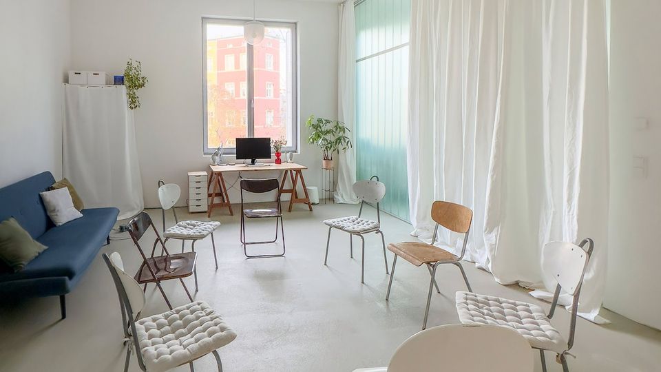 Heller, minimalistischer Raum mit weißem Boden und hohen Decken, mehreren verstreut stehenden Stühlen und einem Tischarbeitsplatz am Fenster. Rechts befindet sich eine große Glasfront mit weißen Vorhängen und grünlich getöntem Licht; links stehen ein blaues Sofa und ein hoher Schrank.