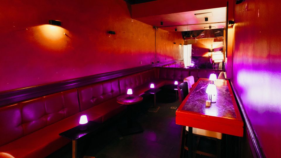 Schmaler Lounge- oder Barbereich mit roten Wänden und durchgehender, gepolsterter Sitzbank entlang der linken Seite. Kleine Tische mit violett leuchtenden Tischlampen und ein langer Hochtisch rechts führen zu einem Spiegel am Ende des Raums.