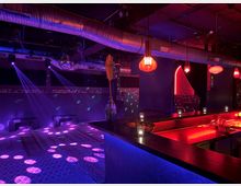 Ein moderner Nachtclub mit einer Bar im Vordergrund, beleuchtet von roten und blauen Lichtern. Im Hintergrund befinden sich Sitzbänke, eine Discokugel und Lichtstrahlen, die punktförmige Muster auf den Boden projizieren; die Decke zeigt sichtbare Lüftungsrohre.