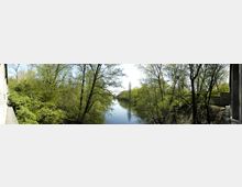 Flusslandschaft mit dichtem Baumbewuchs an beiden Ufern, die frühlingshaft grün sind, und einem klaren, ruhigen Fluss in der Mitte. Im Hintergrund ist eine kleine Brücke zu erkennen, die den Fluss überspannt.