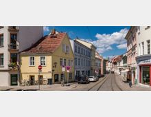 Straßenszene in einer Altstadt mit gepflasterten Straßen, Straßenbahnschienen und mehrstöckigen Gebäuden mit farbigen Fassaden. Ein gelbes Haus mit rotem Ziegeldach steht im Vordergrund, während Autos und Fahrräder am Straßenrand geparkt sind.