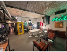 Ein Raum mit Gewölbedecke und freiliegenden weißen Ziegelwänden, eingerichtet im Retro-Stil. Zu sehen sind eine gelbe Telefonzelle, Neon-Schriftzüge, Designermöbel mit geschwungenen Formen sowie Regale und Lampen im Vintage-Look.