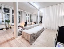 Heller Raum mit holzfarbenem Boden, weißen Wänden und großen Fenstern mit Sprossen, die von langen Vorhängen umrahmt sind. Im Vordergrund stehen mit Tüchern bedeckte Möbel, im Hintergrund sind weitere Sitzgelegenheiten sowie Pflanzen und Dekorationen sichtbar.