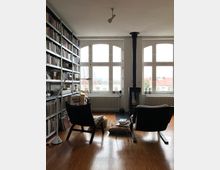 Ein Wohnzimmer mit großen, weißen Fenstern, einem freistehenden Kaminofen und einem Holzboden. Links stehen hohe Bücherregale, rechts befinden sich zwei Sessel mit Kissen und Büchern, daneben ein kleiner Beistelltisch.