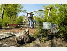 Ein Spielplatz mit Holzgeräten, darunter eine Rutsche und eine Schaukel, umgeben von Bäumen und Büschen. Im Vordergrund steht eine Informationstafel am Zaun sowie ein großer Stein, neben einem gepflasterten Weg.
