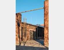 Backsteingebäude eines historischen Industriekomplexes mit großen Metalltüren, Gitterfenstern und angrenzenden Schornsteinen. Im Vordergrund ist eine Leiter an der Wand befestigt, und das gepflasterte Gelände ist von Schatten durchzogen.