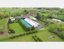 Luftaufnahme eines landwirtschaftlichen Grundstücks mit mehreren Gewächshäusern, einem Hauptgebäude und einer Wiese, umgeben von Bäumen und Feldern. Im Vordergrund gibt es einen kleinen Lagerbereich und eine Feuerstelle, im Hintergrund stehen weitere Gebäude und eine weitläufige grüne Landschaft.