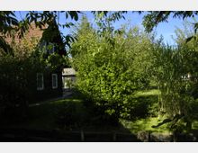 Ein ländlicher Garten mit dichter Vegetation, darunter Bäume, Sträucher und eine Wiese, grenzt an ein dunkles Holzhaus mit roten Dachziegeln. Im Hintergrund sind weitere Gebäude und Pflanzen erkennbar, die Szene ist von natürlichem Licht durchflutet.