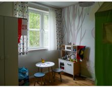 Ein Kinderzimmer mit einem großen Fenster, das von gemusterten Vorhängen umrahmt ist, und einer Tapete mit Baum- und Heißluftballon-Motiven. Es gibt eine kleine Spielküche, einen runden Tisch mit Hockern sowie Spielzeug auf dem Boden und in einem Regal.