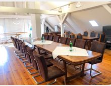 Heller Dachgeschossraum mit einem langen Holztisch, umgeben von braunen Stühlen mit Metallfüßen. Im Hintergrund befinden sich weitere Sitzgruppen, Holzfußboden und sichtbare Dachschrägen mit Dachfenstern.