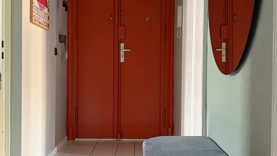 Narrow apartment entry hallway with light gray walls and white tiled floor, leading to a large red double door with matching built-in cabinets above. A round wall mirror and a cushioned bench sit along the right wall, with a framed poster on the left and doorways opening off the corridor.