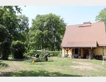 Ein Einfamilienhaus mit gelber Fassade und rotem Satteldach steht in einem großen Garten mit Rasenfläche, Bäumen und Büschen. Im Vordergrund liegen Gartenmöbel und Spielzeug, während der Hintergrund von dichter Vegetation und Bäumen geprägt ist.