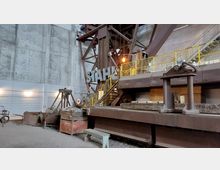 Industrielle Innenansicht einer ehemaligen Stahlproduktionsanlage mit großen Metallstrukturen, Geländern und Treppen. Im Vordergrund stehen Maschinen, Metallträger und das Wort "STAHL" als dekoratives Element auf der Anlage.