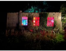 Backsteinfassade eines niedrigen Industriegebäudes bei Nacht mit drei großen, rundbogigen Sprossenfenstern, die innen in kräftigem Blau und Rot beleuchtet sind. Efeu und dichtes Gartenbewuchs umrahmen das Gebäude, im Hintergrund stehen hohe Bäume vor dunklem Himmel.