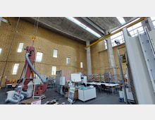 Großer, industrieller Innenraum mit hohen Wänden aus gelblich gemusterten Ziegeln, Oberlichtern und Betonstützen. Der Raum enthält technische Geräte, Tische mit Stühlen und eine rote Maschine mit Schläuchen, die in der Mitte prominent platziert ist.