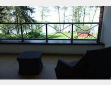 Blick von einer Terrasse mit gefliestem Boden und schwarzen Rattanmöbeln auf eine grüne Wiese, umgeben von Bäumen, mit einem See und einem kleinen roten Gebäude im Hintergrund. Ein schmiedeeisernes Geländer rahmt den Ausblick ein.