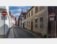 Eine schmale gepflasterte Straße mit teils sanierten, teils verfallenen Häusern; rechts ein Gebäude mit zugenagelten Fenstern, links helle Fassaden. Im Hintergrund ist eine Kirche mit rotem Ziegeldach und einem Turm sichtbar. Schilder markieren die Straße als Einbahnstraße, Fahrräder sind jedoch erlaubt.
