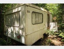 Ein älterer, leicht verwitterter Wohnwagen steht in einem bewaldeten Gebiet. Der Wohnwagen hat ein kleines Fenster mit Gardine, ein offenes Eingangstor und ist von dichtem Grün umgeben.