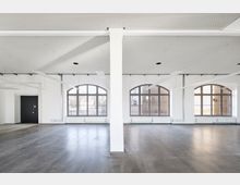 Großer, leerer Innenraum mit einem modernen, grauen Boden, weißen Wänden und einer Lochdecke mit eingelassenen Leuchten. Große Bogenfenster lassen viel Tageslicht in den Raum, und eine weiße Säule steht mittig im Vordergrund.