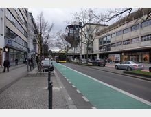 Straßenszene in einer Einkaufsstraße in einer Stadt, mit modernen Gebäuden, Geschäften und Schaufenstern auf beiden Seiten. In der Mitte der Straße befinden sich ein zweispuriger Fahrweg, ein grüner Fahrradweg, sowie ein vorbeifahrender gelber Stadtbus; mehrere Fußgänger und parkende Autos sind sichtbar.