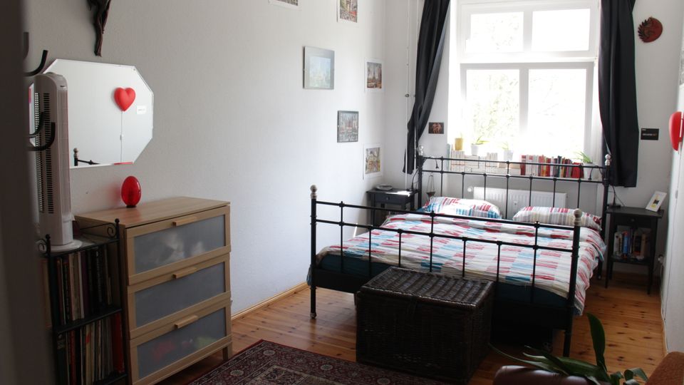 Bright bedroom with a black metal-framed bed and patterned duvet, set on a wooden floor with a rug and a storage bench at the foot. A large window with dark curtains and a low bookshelf along the sill faces the bed, with a dresser and shelving against the left wall.